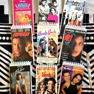 May include: A collection of eight spiral-bound notebooks featuring covers with movie posters for The Little Mermaid, Pretty in Pink, Backstreet Boys, Speed, Uncle Buck, The Net, Flashdance, Hook, and She's All That, arranged in a grid pattern.