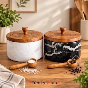 May include: Two cylindrical containers with white and black marble bodies and circular wooden lids featuring a central knob. Each container includes a small wooden scoop, one filled with white salt crystals and the other with black peppercorns.
