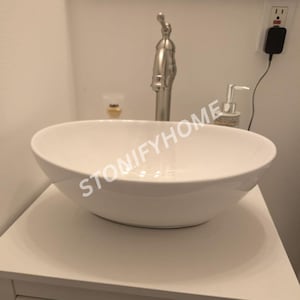 May include: A white oval ceramic vessel sink with a smooth, glossy finish. The basin is positioned on a white vanity surface. A tall, brushed nickel faucet stands behind the sink. The text STONIFYHOME is overlaid across the center of the basin.