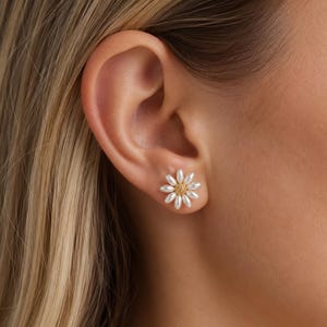 Puede incluir: Un pendiente de botón con forma de flor de margarita, que presenta pétalos ovalados de perla sintética blanca dispuestos en un patrón circular alrededor de un pequeño centro metálico texturizado de tono dorado. Se sujeta al lóbulo con un perno.