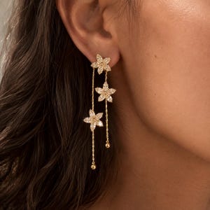 Puede incluir: Un pendiente colgante de tono dorado con tres dijes en forma de flor incrustados con piedras transparentes brillantes. El diseño incluye dos cadenas delicadas de diferentes longitudes, cada una terminada en una pequeña cuenta dorada.