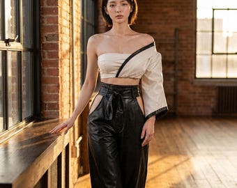 May include: A two-piece outfit consisting of an off-the-shoulder cream-colored crop top with a single long sleeve and black trim, paired with high-waisted black leather trousers featuring side cutouts and a tie-waist belt detail.