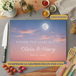 May include: A rectangular tempered glass cutting board featuring a sunset sky with a full moon, the text "UNDER THE SAME MOON", the names "Olivia & Henry", and the locations "Boston - Seoul". The board is printed in the USA.