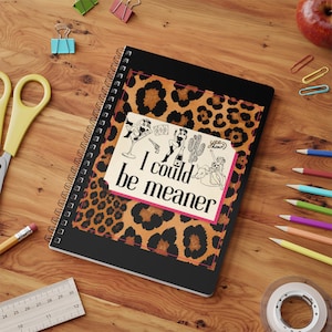 May include: A spiral-bound notebook with a black cover featuring a central leopard print panel. The panel displays the text "I could be meaner" alongside illustrations of a person in a martini glass, a revolver, a cactus, and a person riding a bull.