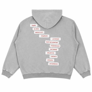 Puede incluir: Una sudadera con capucha gris que presenta una serie de parches rectangulares blancos con bordes sin rematar, dispuestos en una columna vertical escalonada en la parte posterior. Cada parche muestra el nombre de una ciudad en texto rojo: Tampa, El Paso, Mexico City, Stamford, Las Vegas, East Rutherford, Foxborough, Baltimore, Arlington, Toronto, Chicago y Los Angeles.