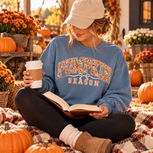 Puede incluir: Una sudadera azul de cuello redondo con el texto PUMPKIN SEASON en una fuente con motivos florales y un lazo, combinada con una gorra beige, leggings negros y botas marrones, mientras sostiene un vaso de papel blanco y un libro abierto.