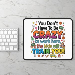 May include: A rectangular white mousepad with a black border featuring the text "You Don't Have To Be CRAZY to work here, the kids will TRAIN YOU!" surrounded by colorful illustrations of school supplies, sports balls, and a group of children.