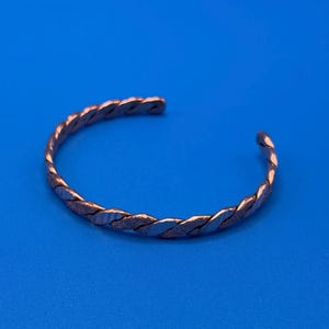 May include: A copper-coloured metal cuff bracelet featuring a twisted rope design. The surface of the bracelet alternates between smooth, reflective sections and textured, matte segments, forming a continuous braided pattern around the open band.