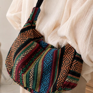 May include: A hobo-style shoulder bag featuring a vibrant woven textile pattern with geometric shapes and stripes in orange, red, green, blue, and black. The bag has a wide, integrated fabric strap and a soft, slouchy construction with a textured finish.