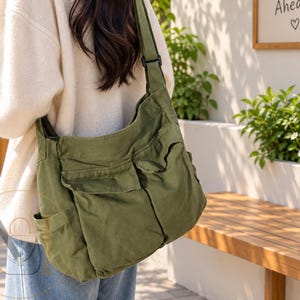 May include: An olive green canvas shoulder bag featuring a wide adjustable strap, two large front pockets with flap closures, and side pockets. The bag has a soft, slouchy structure and is worn over the shoulder, displaying a utilitarian design.