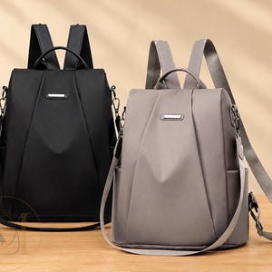 May include: Two backpacks, one in black and one in gray, featuring a structured design with a top handle, adjustable shoulder straps, side pockets, and a central rectangular metal accent on the front flap. Both bags have a smooth finish.