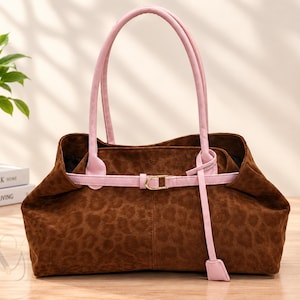 May include: A brown leopard print handbag featuring a soft suede-like texture, two light pink shoulder straps, and a horizontal light pink belt detail across the front with a gold-toned buckle and a dangling light pink strap end.