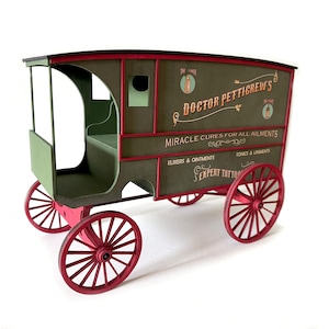 Op de afbeelding: Een decoratief houten model van een vintage wagen met olijfgroene panelen, rode randen en felrode spaakwielen. Op de zijkant staat de tekst Doctor Petticrew's, Miracle Cures For All Ailments, Elixirs & Ointments, Tonics & Liniments.