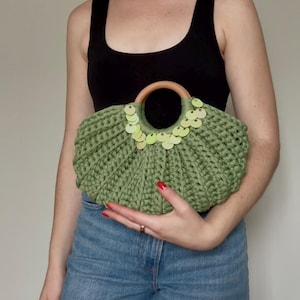 May include: A sage green crocheted handbag featuring a semi-circular silhouette and a round wooden handle. The bag is embellished with a row of light green circular sequins arranged along the top edge, surrounding the wooden handle attachment.