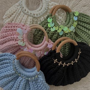 Peut inclure: Cinq sacs à main crochetés avec des anses rondes en bois, déclinés en blanc, vert sauge, rose clair, bleu clair et noir. Le sac blanc présente des perles, les sacs vert et rose ont des sequins irisés, et le sac noir porte des breloques abeilles.