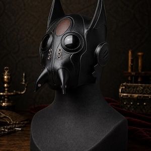May include: A black leather mask featuring two pointed ears, two large circular dark lenses, and two downward-pointing conical protrusions at the nose area. The mask includes a brown oval patch on the forehead and metallic eyelet details.
