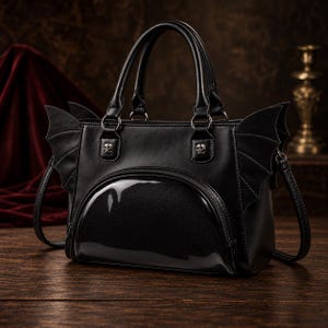 Puede incluir: Un bolso de mano negro con paneles laterales en forma de alas de murciélago, dos asas superiores y un bolsillo frontal semicircular con ventana transparente. El bolso incluye dos tachuelas metálicas plateadas con forma de calavera.