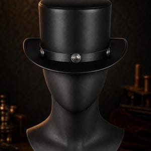 Può includere: Un cilindro nero con una finitura liscia e opaca e una corona strutturata. Una fascia in pelle nera circonda la base della corona, decorata con tre inserti metallici circolari color argento caratterizzati da dettagli in rilievo.