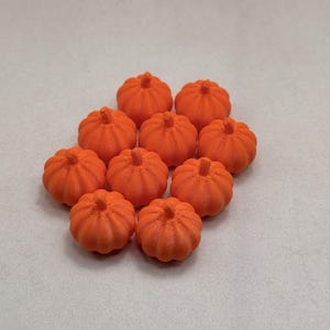 May include: A collection of ten small, bright orange, three-dimensional pumpkin-shaped objects. Each pumpkin features a ribbed texture and a small, centered stem at the top. The items are arranged in a cluster, displaying a uniform matte finish.
