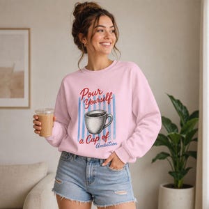 Pode incluir: Uma camisola de manga comprida cor-de-rosa claro com um gr&aacute;fico de uma ch&aacute;vena de caf&eacute; centrado sobre riscas verticais azuis. O texto "Pour Yourself a Cup of Ambition" est&aacute; impresso em letras vermelhas e azuis &agrave; volta da ilustra&ccedil;&atilde;o.