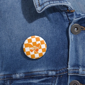 May include: A circular pin-back button featuring an orange and white checkered pattern. Centered on the button is a solid orange butterfly silhouette overlaid with a white cursive script text that appears to read as a stylized signature or logo.