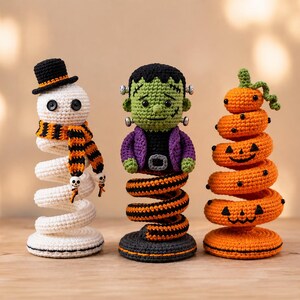 May include: Three crocheted Halloween figures on spring bases. The left is a white snowman with a black hat and orange-black scarf. The center is a green Frankenstein monster with a purple jacket. The right is a stack of orange jack-o'-lanterns.