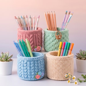 May include: Four cylindrical crochet pen holders in pink, mint green, light blue, and beige. Each holder features a textured knit pattern and small floral embellishments. The holders are filled with various pens, pencils, and brushes.