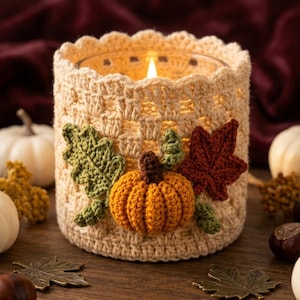 May include: A cylindrical glass candle holder encased in a cream-colored crocheted cozy. The cozy features a textured lattice pattern and is decorated with a small orange crocheted pumpkin, a green crocheted leaf, and a dark red crocheted leaf.