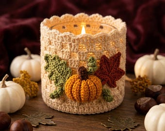 May include: A cylindrical glass candle holder encased in a cream-colored crocheted cozy. The cozy features a textured lattice pattern and is decorated with a small orange crocheted pumpkin, a green crocheted leaf, and a dark red crocheted leaf.