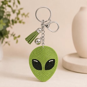 Op de afbeelding: Een sleutelhanger met een groene alien-hoofd bedel bedekt met kleine strass-steentjes en grote zwarte strass-ogen. De bedel is bevestigd aan een zilverkleurige metalen ring en karabijnhaak, vergezeld door een groen kwastje en een zilveren metalen kraal.