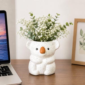 Puede incluir: Una maceta de cerámica blanca moteada con forma de koala, que presenta una nariz marrón, ojos negros y orejas redondeadas. La maceta contiene un ramo de pequeñas flores blancas y hojas de eucalipto verdes como arreglo decorativo.