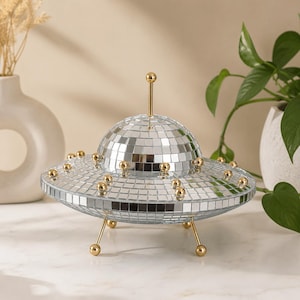 May include: A decorative UFO-shaped object covered in small square mirror tiles, featuring a central dome and a wide disc base. The piece is accented with gold-colored metal spheres along the rim and top, and stands on three thin gold-colored metal legs.