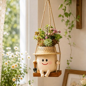 May include: A hanging planter shaped like a cream-colored character with a smiling face, wearing a straw hat, and sitting on a small wooden swing. The planter holds a variety of green succulents with small pink flowers and includes a tiny shovel.