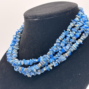 May include: A multi-strand necklace composed of numerous small, irregular, tumbled blue lapis lazuli stone chips. The necklace features several layered strands of these polished, vibrant blue gemstone fragments, creating a textured appearance.