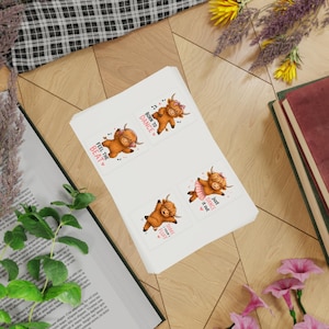 Op de afbeelding: Een vel met vier vierkante stickers met cartoon Highland-koeien. Eén koe draagt een koptelefoon met de tekst "FEEL THE BEAT", een andere een strik met "BORN TO DANCE", één met een tutu "JUST DANCE IT OUT" en één met "SHAKE YOUR FLUFF".