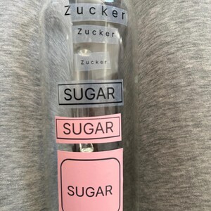 Könnte beinhalten: Ein durchsichtiger Glasbehälter mit mehreren Etiketten, die den Text Zucker in verschiedenen Größen zeigen, gefolgt von dem Wort SUGAR in einem schwarz umrandeten Kasten und zwei weiteren Etiketten mit dem Wort SUGAR auf rosa Hintergrund.