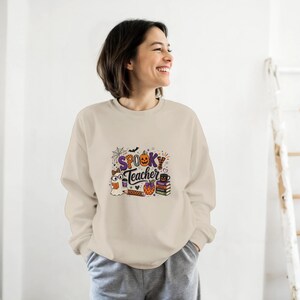 Può includere: Una felpa beige a maniche lunghe con una stampa grafica che riporta il testo "SPOOKY Teacher" in viola e arancione. Il design include un fantasma, una zucca, una pila di libri, una matita e una tazza di caffè con motivi di ragnatele e pipistrelli.