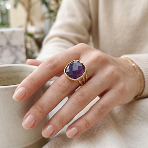 May include: A gold ring featuring a large, square-cut, faceted purple amethyst gemstone set in a textured gold bezel. The ring band consists of three thin, twisted gold wires that converge into a single band at the base of the finger.