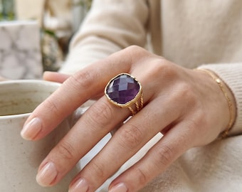 May include: A gold ring featuring a large, square-cut, faceted purple amethyst gemstone set in a textured gold bezel. The ring band consists of three thin, twisted gold wires that converge into a single band at the base of the finger.