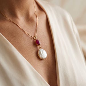 Könnte beinhalten: Eine goldfarbene Halskette mit einer feinen Kette und einem Anhänger, der aus einem kleinen, facettierten, dunkelrosa Edelstein über einer größeren, unregelmäßigen, weißen Barockperle besteht, beide in goldfarbenen Metallfassungen.