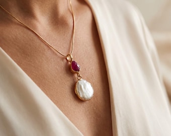 May include: A gold-toned necklace featuring a delicate chain with a pendant consisting of a small, faceted, deep pink gemstone positioned above a larger, irregular, white baroque pearl, both set in gold-toned metal frames.