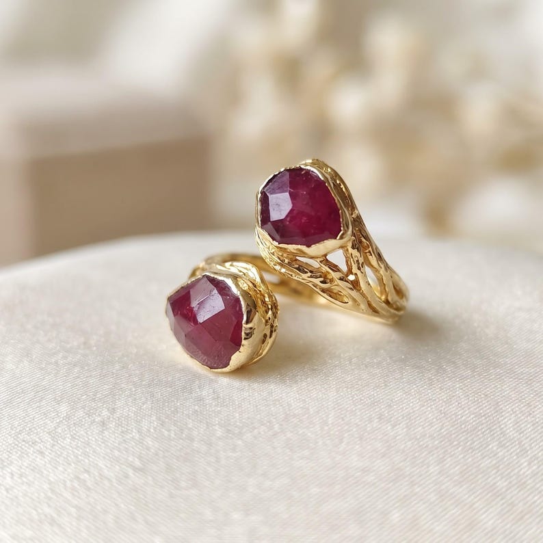 May include: A gold open ring featuring two faceted, deep red ruby gemstones. The band has a textured, organic, branch-like appearance, with one ruby set at each end of the open design, creating a bypass style ring with a polished finish.