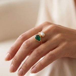 May include: A gold open-style ring featuring a teardrop-shaped green gemstone on one end and a small, round white pearl on the other. The band is thin and polished, showcasing a contrasting design between the vibrant green stone and the smooth pearl.