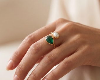 May include: A gold open-style ring featuring a teardrop-shaped green gemstone on one end and a small, round white pearl on the other. The band is thin and polished, showcasing a contrasting design between the vibrant green stone and the smooth pearl.