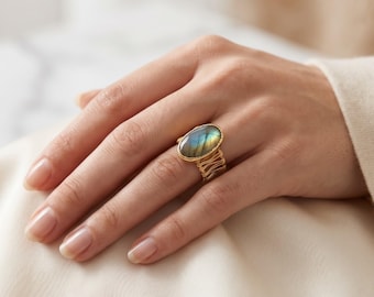 May include: A gold ring featuring an oval-shaped labradorite gemstone with blue and yellow iridescence. The band is constructed from multiple thin gold wires wrapped around the finger, creating a textured, layered appearance.