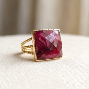 May include: A gold ring featuring a large, square-cut, faceted deep pink gemstone set in a gold frame. The band of the ring is composed of three thin, textured gold wires that converge into a single shank at the back of the ring.