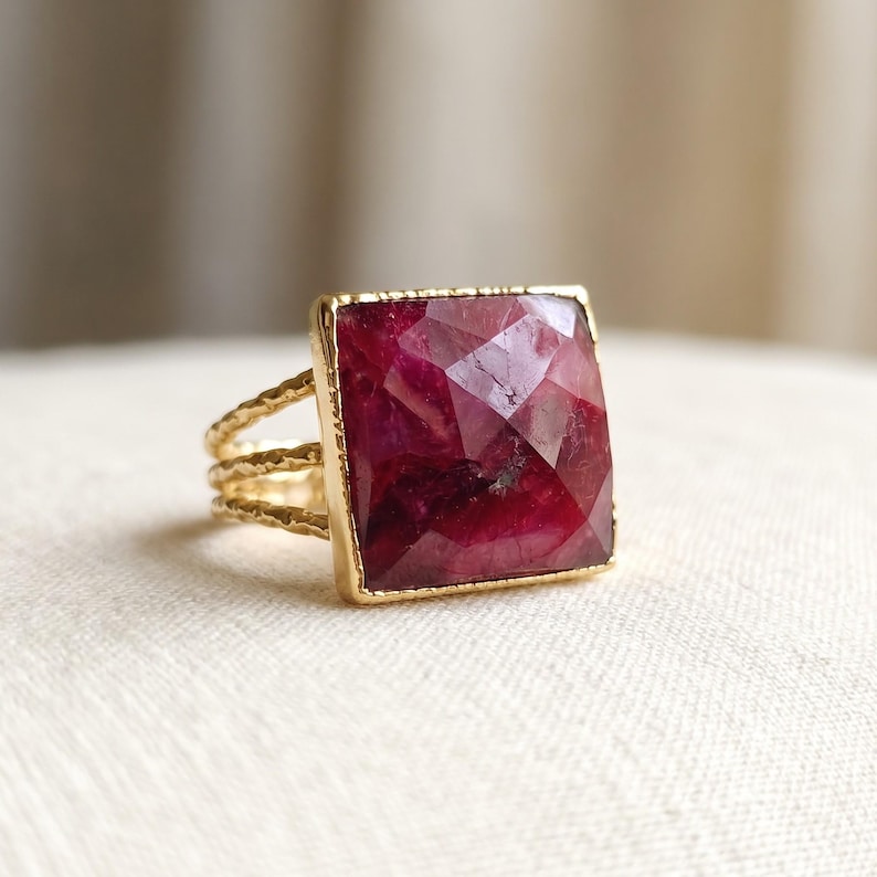 May include: A gold ring featuring a large, square-cut, faceted deep pink gemstone set in a gold frame. The band of the ring is composed of three thin, textured gold wires that converge into a single shank at the back of the ring.