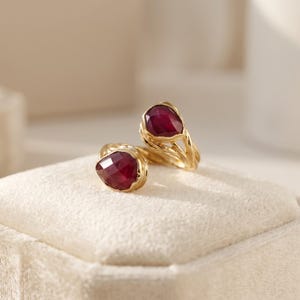 May include: A gold bypass ring featuring two faceted, pear-shaped deep red gemstones. The band has a textured, organic appearance with intertwined metal strands that hold each stone securely at the open ends of the ring design.