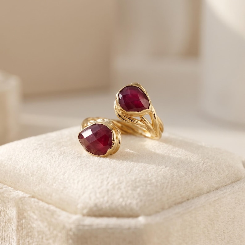 May include: A gold bypass ring featuring two faceted, pear-shaped deep red gemstones. The band has a textured, organic appearance with intertwined metal strands that hold each stone securely at the open ends of the ring design.