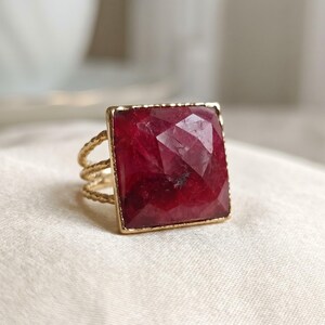 May include: A gold ring featuring a large, square, faceted deep red gemstone set in a thin, metallic frame. The band of the ring is constructed from three delicate, twisted gold wires that converge into a single shank at the base.